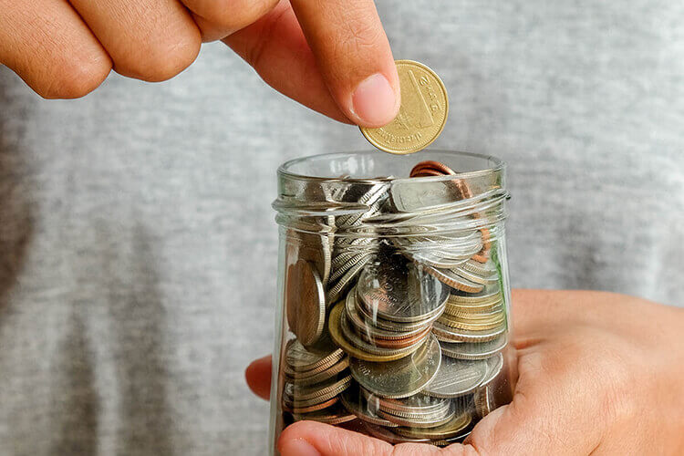 A person adds a coin to a jar brimming with coins, illustrating the importance of saving and building wealth.