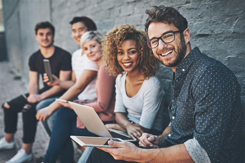 A group of diverse, smiling milennials sitting on a bench in front of a grey brick wall.